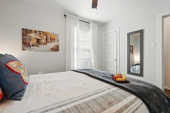 Un dormitorio con una cama, un cuadro, una ventana con cortinas, un espejo y una puerta.