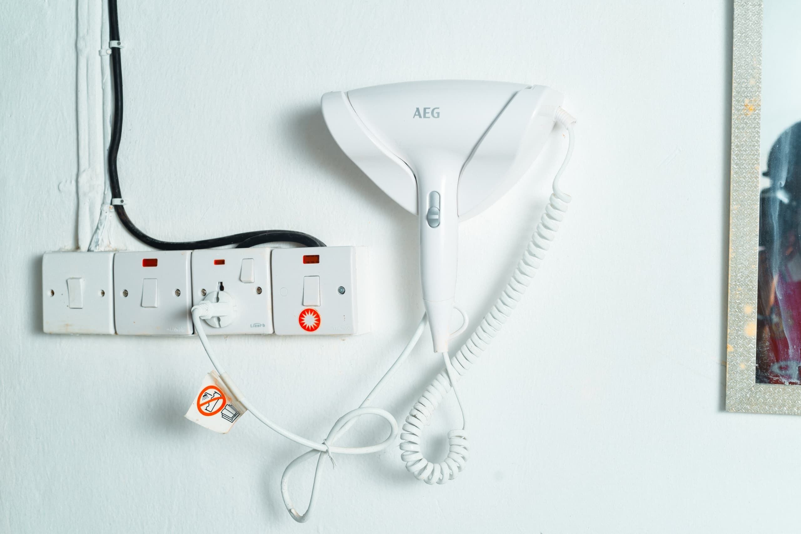 A white AEG hairdryer connected to a wall socket with a power cord.