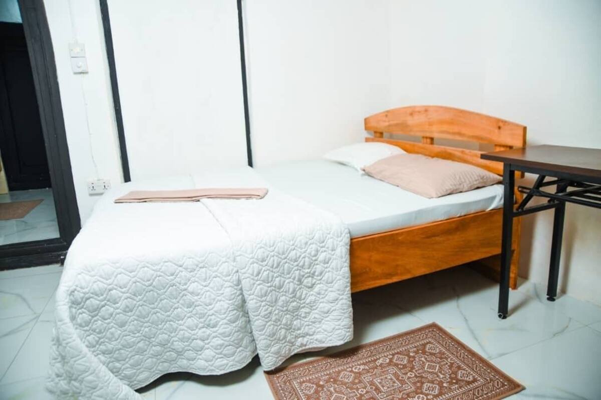 A single bed with a wooden headboard, a bedside table, and a patterned rug on a tiled floor.