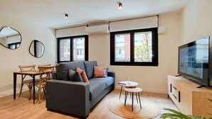 Una sala moderna con un sofá gris, una mesa de comedor de madera y sillas, un televisor sobre un mueble de madera y ventanas grandes con persianas.