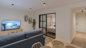 Una sala moderna con una televisión de pantalla plana, un sofá gris, una mesa de comedor con sillas y un ventanal corredero que da a un dormitorio.