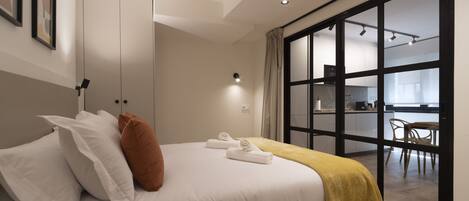 A modern bedroom with a bed, white bedding, a brown pillow, and a yellow throw blanket. There is a large window with a view of a kitchen and dining area.