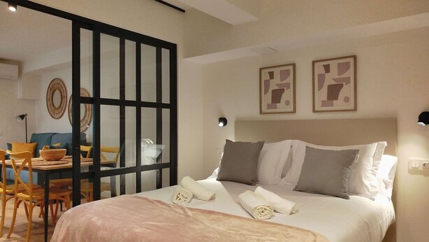 Une chambre bien éclairée, avec un lit, des oreillers et une table de chevet. On aperçoit un coin repas avec des chaises et une table à travers une cloison en verre.