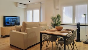 Una sala de estar moderna con un sofá, una mesa de comedor con sillas, una planta en maceta y un televisor sobre un mueble.
