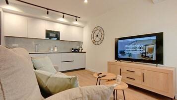 A modern living room with a sofa, a television displaying a webpage, a kitchen area, and a clock on the wall.