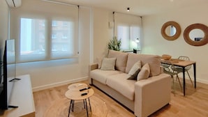 Una sala moderna con un sofá color beige, una mesita redonda pequeña y una mesa de comedor con sillas. Hay un ventanal grande que proporciona luz natural, y dos espejos colgados en la pared.
