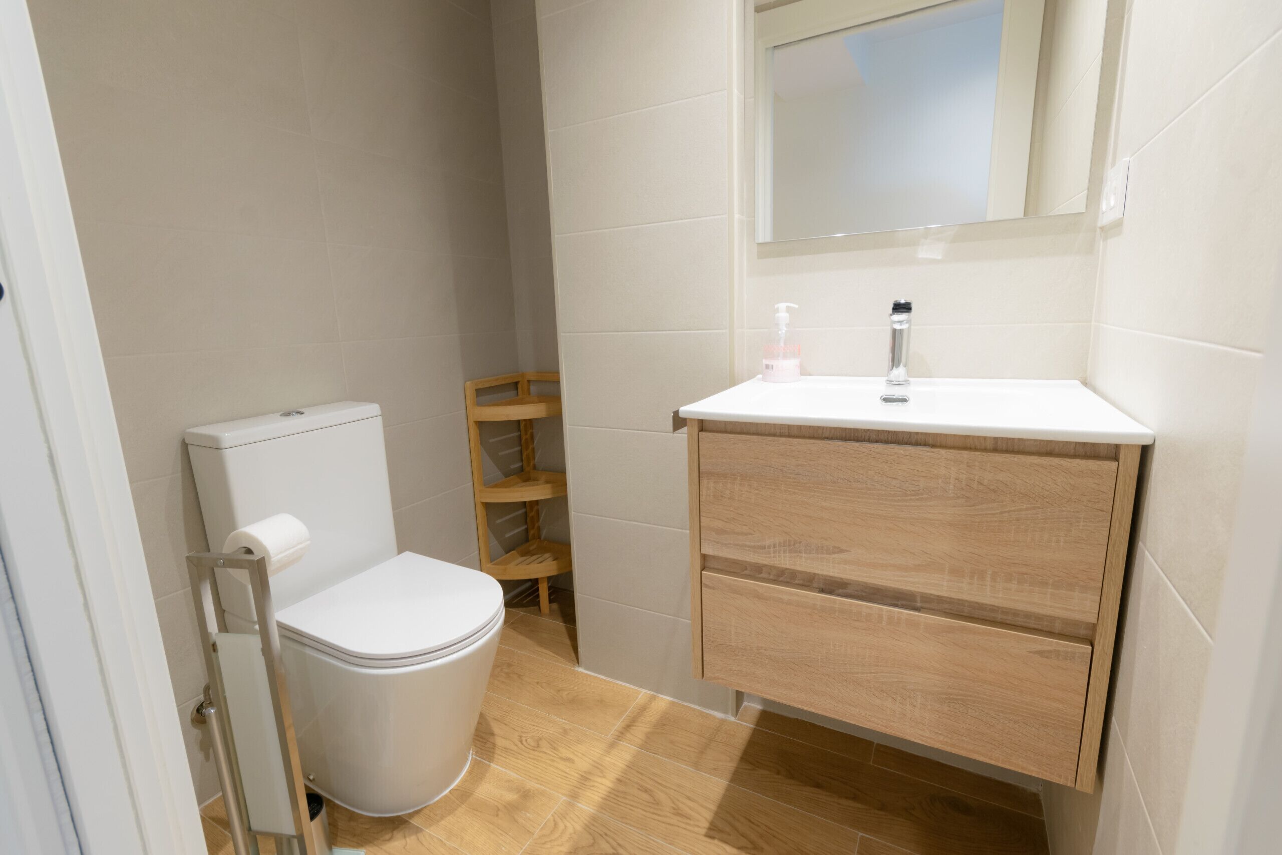 Et lille badeværelse med toilet, en træhylde, en hvid vask med spejl og en hvid toiletpapirsholder.