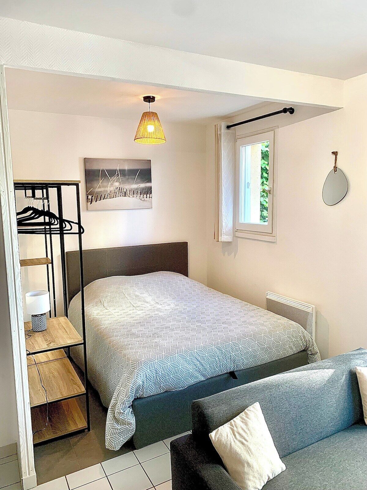 Un dormitorio con cama, mesita de noche, lámpara, lámpara colgante, ventana con cortinas, espejo adosado a la pared y un cuadro enmarco.