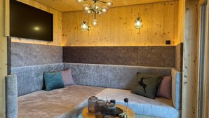 Ein gemütliches Zimmer mit Holzbalkendecke, einem großen grauen Sofa, einem kleinen runden Tisch mit Dekoration und einer Deckenleuchte.