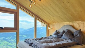 Ein Schlafzimmer mit großem Fenster und Blick auf die Berge, Holzboden und einem Bett mit Kissen.