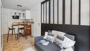 A modern living room with a grey sofa, black and white patterned cushions, and a magazine on the sofa. There is a kitchen area with a white fridge, a microwave, and a wooden countertop with a fruit bowl and a coffee machine. The room has a herringbone wood floor and a large window with blinds. - Modern Central Paris Flat by Les Halles (Paris)