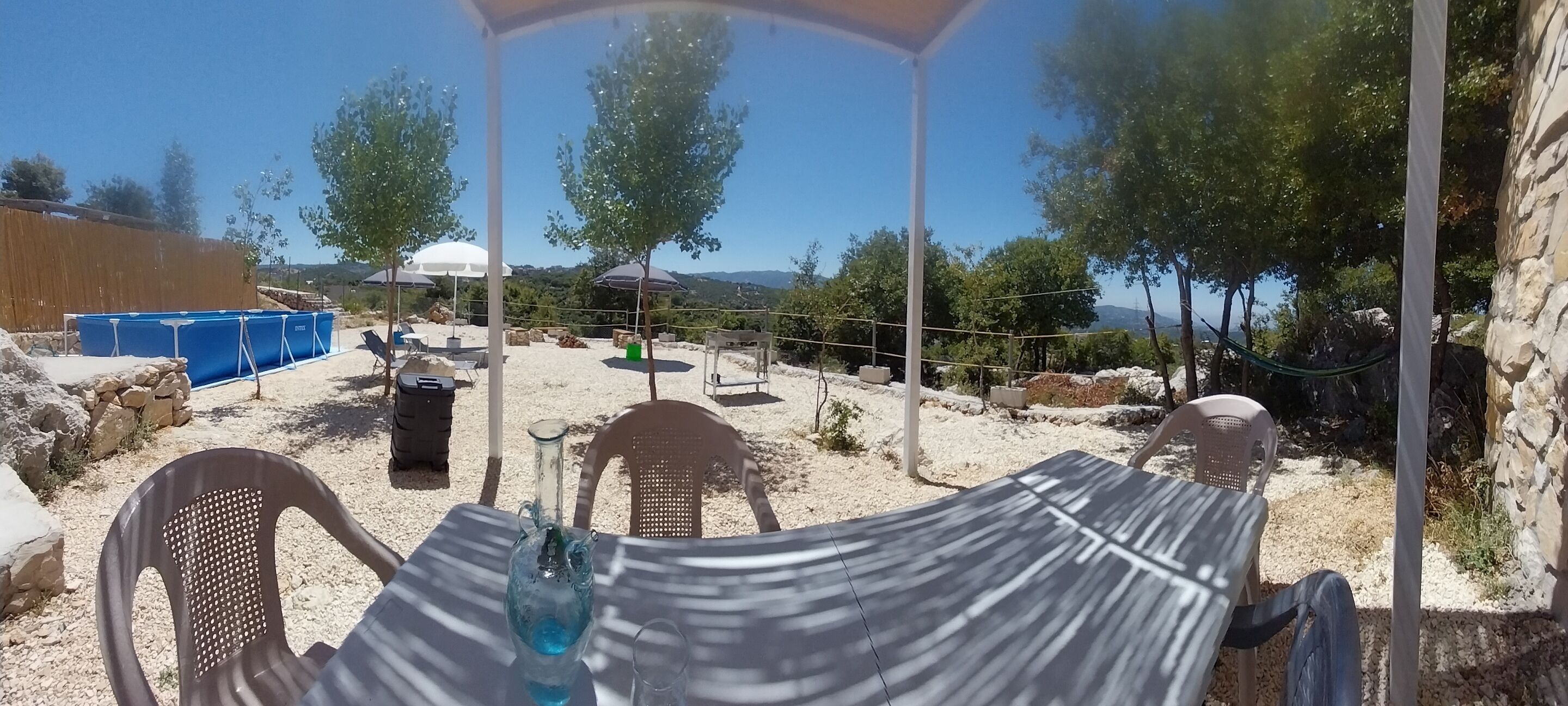 Uma área externa com mesas e cadeiras, uma piscina azul e vista para a paisagem ao redor.