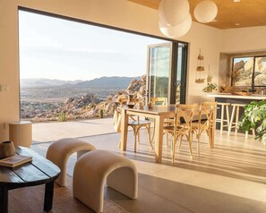 A modern dining area with a large table and chairs, a glass door leading to a patio with a view of the mountains. - Fox Inn • Epic Views, Hot Tub, Cowboy Tub, Grill (Yucca Valley)