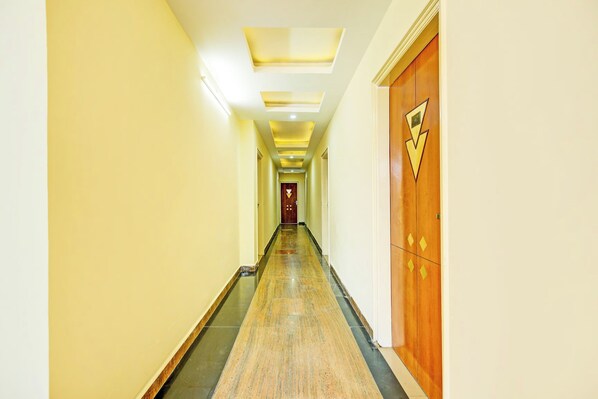 Um corredor comprido e estreito, com uma porta de madeira à direita, piso claro e iluminação embutida no teto.