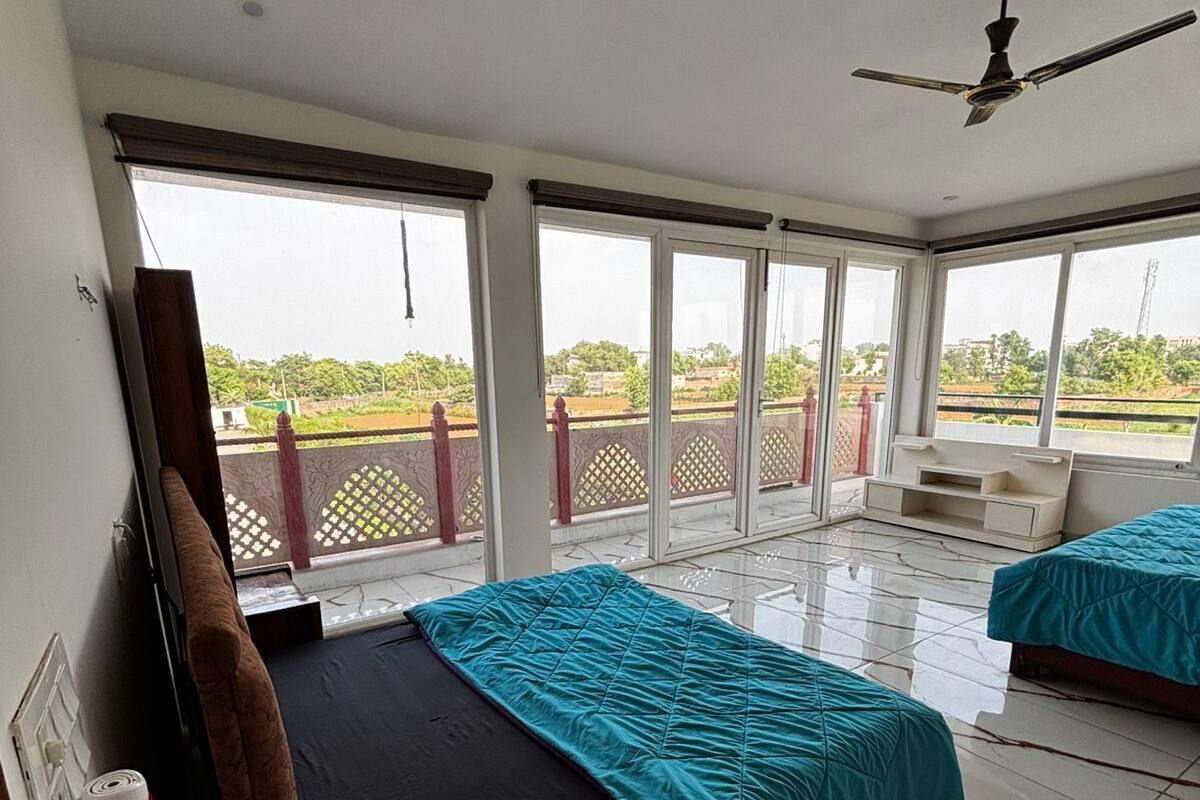 Habitación de hotel con dos camas, ventilador de techo y ventanas amplias con vistas a zonas verdes.
