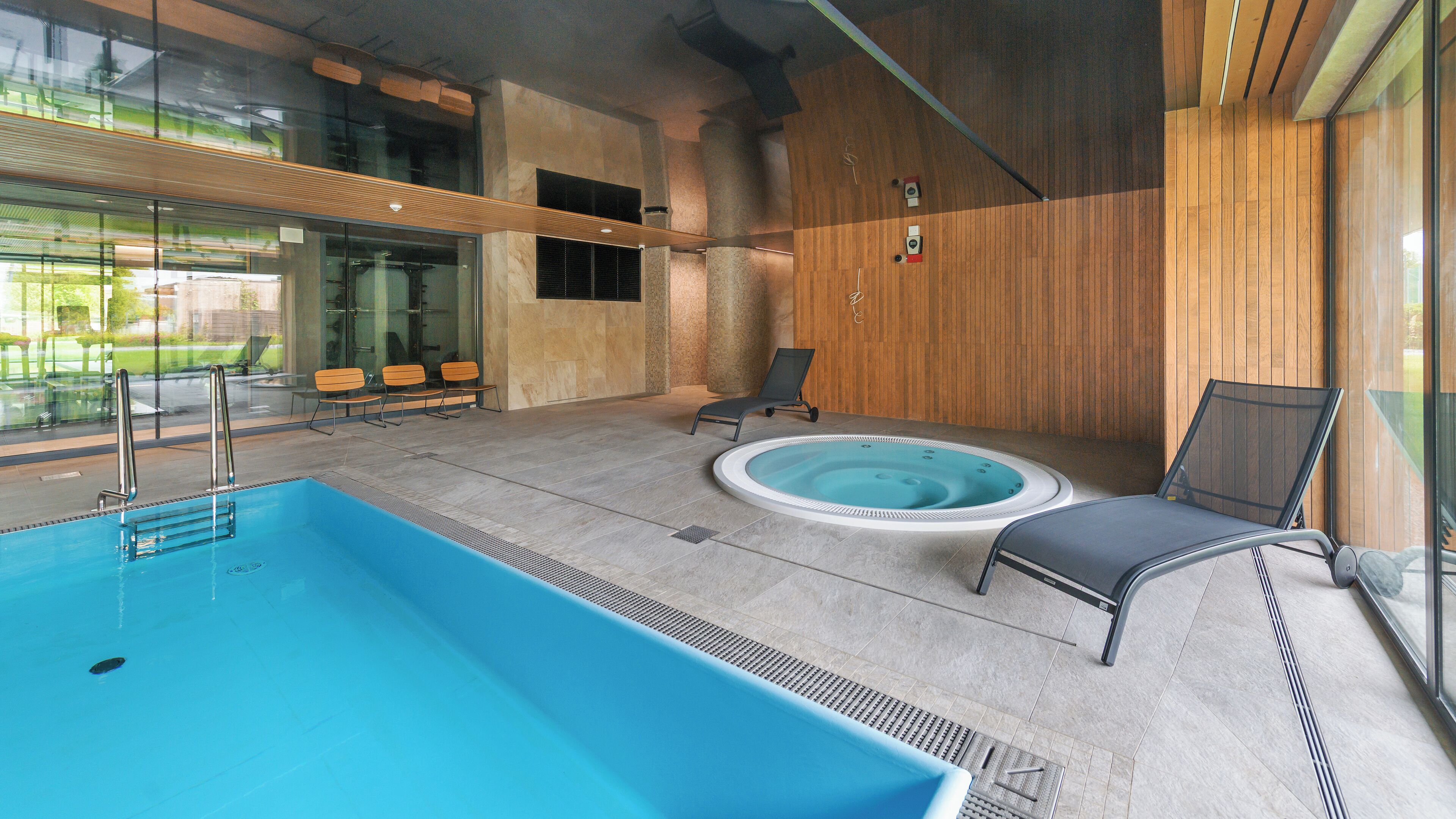 A modern indoor swimming pool area with a hot tub, lounge chairs, and a glass wall offering a view of the outdoors.