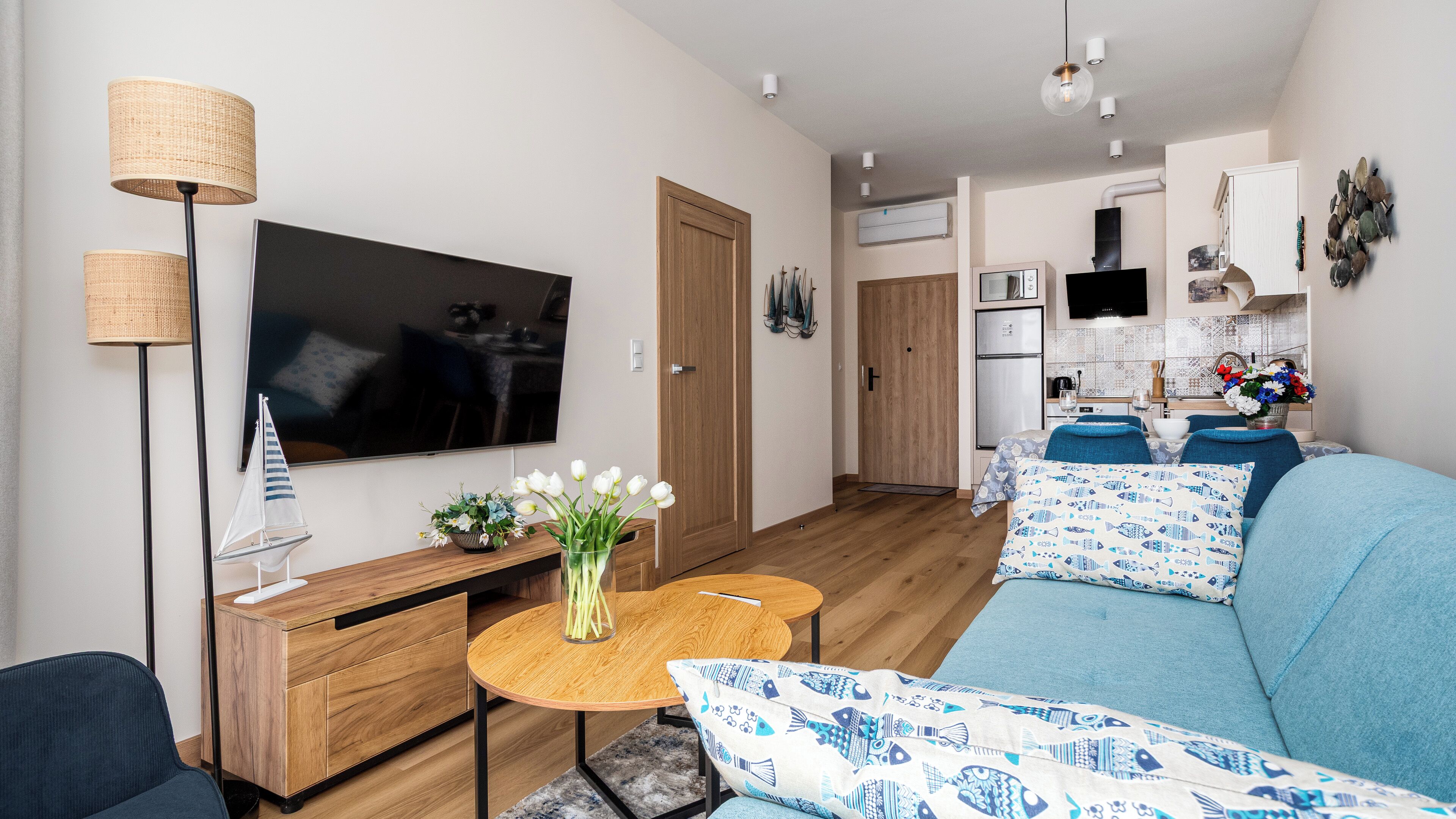 A modern living room with a flat-screen TV, wooden furniture, a small round table with a vase of flowers, and a blue sofa with patterned cushions.