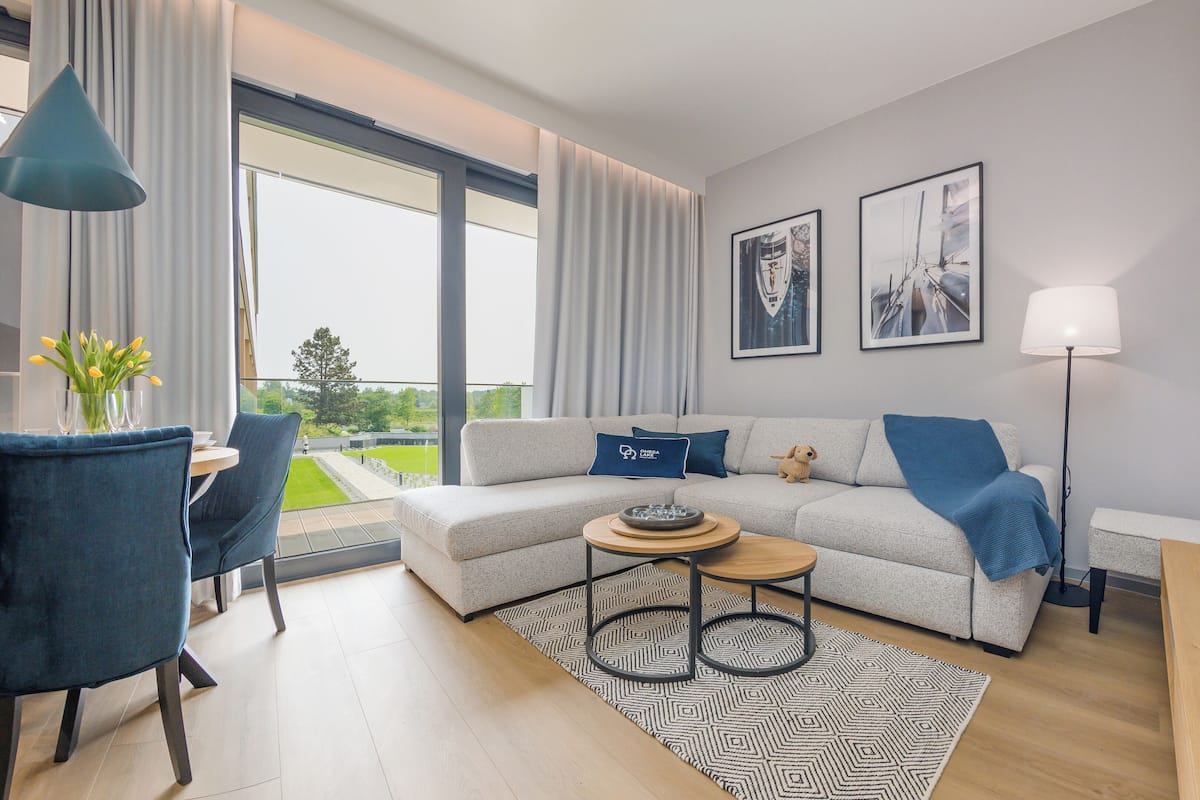 Un salon moderne avec un canapé gris, une table basse et une vue sur un paysage verdoyant grâce à de grandes fenêtres.