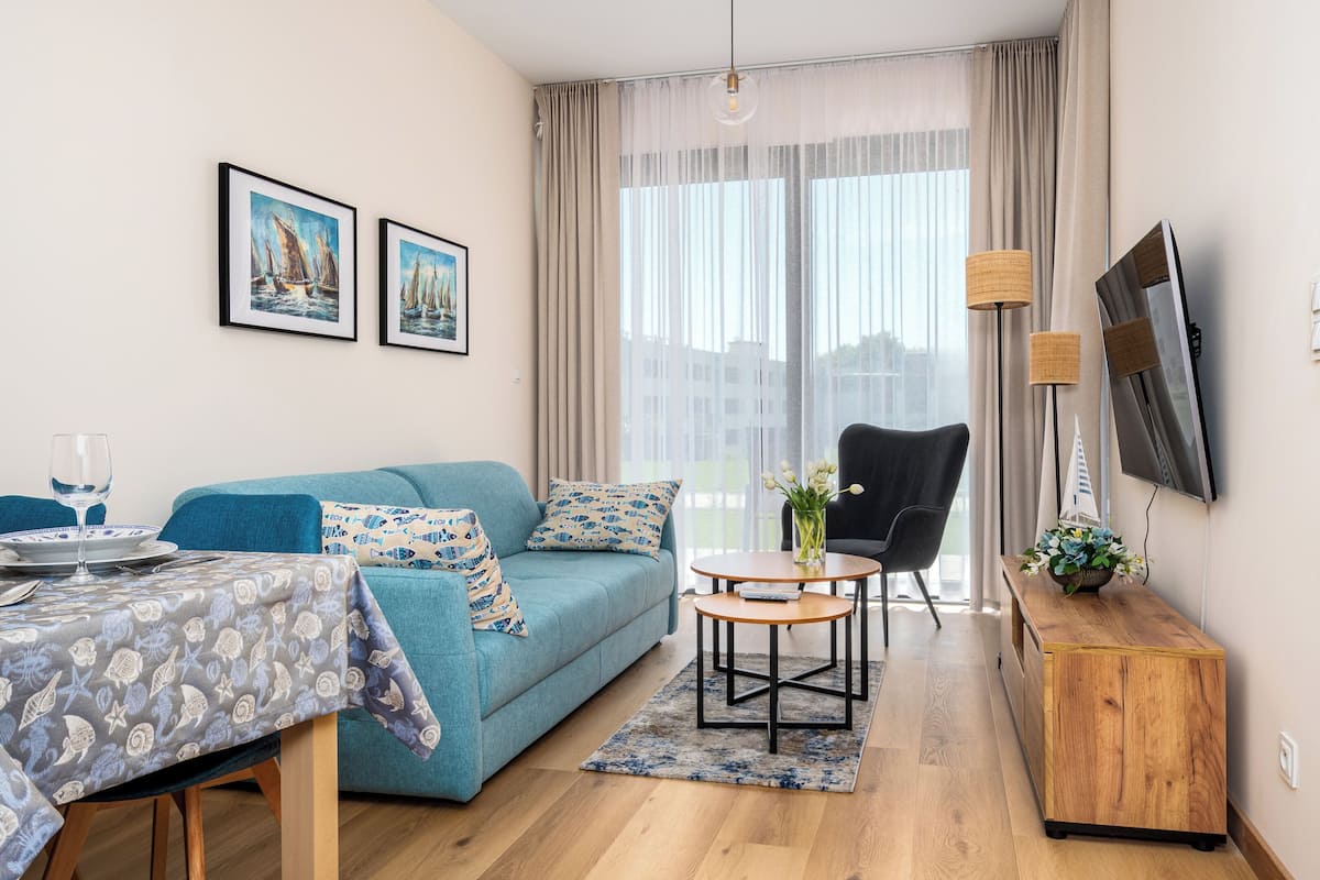 Un salon moderne avec un canapé bleu, une table à manger recouverte d’une nappe à motifs, une petite table ronde ornée d’un vase et un téléviseur fixé au mur.