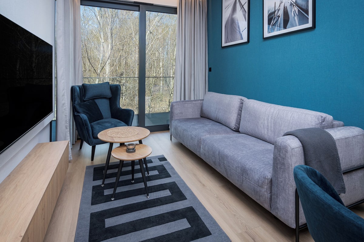 Un salon moderne avec un grand téléviseur, un canapé, un fauteuil, une petite table et une vue sur les arbres par la fenêtre.