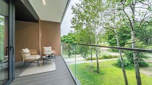 Ein Balkon mit zwei Korbstühlen, einem kleinen Tisch mit Blumen in einer Vase und einem Glasgeländer mit Blick auf eine grüne Landschaft mit Bäumen und einem Weg.