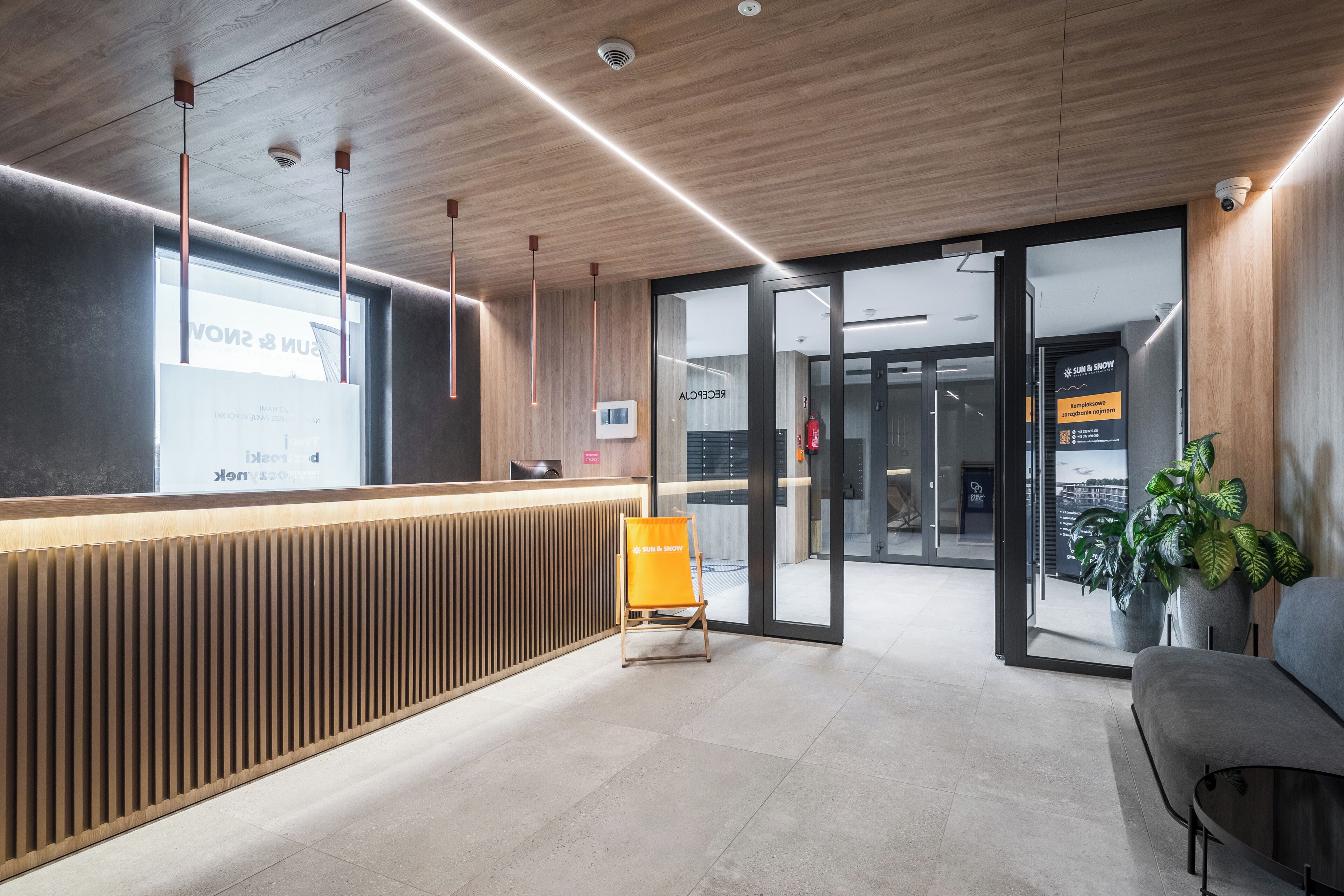 Un espace d’accueil moderne avec un comptoir de réception en bois, un coin salon doté d’un canapé gris et une porte en verre donnant sur une autre pièce.