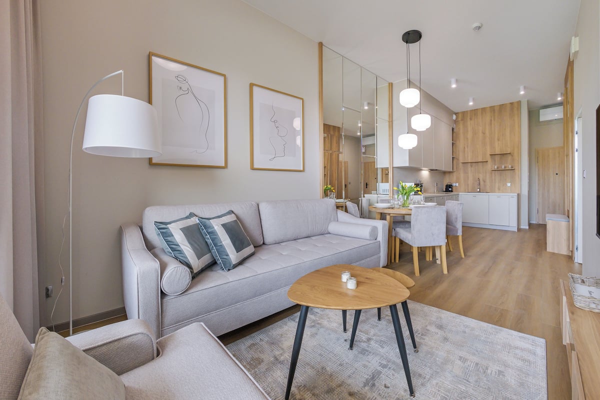 Un salon moderne avec un canapé, une table basse et un coin repas avec des chaises. On aperçoit au fond une cuisine avec des meubles en bois et un évier.