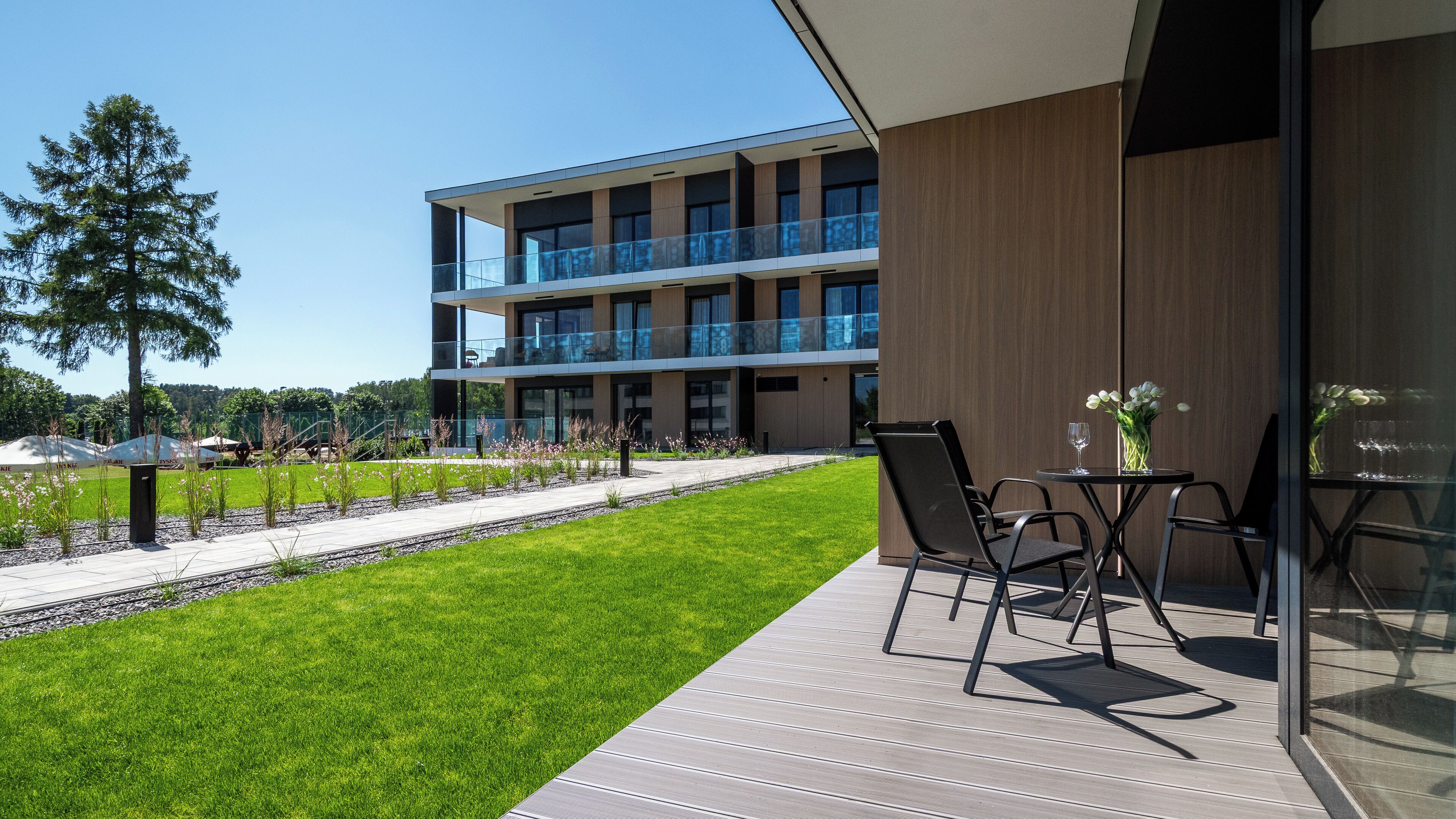 Un bâtiment moderne doté d’un balcon en verre, d’un espace extérieur aménagé pour s’asseoir et d’une pelouse bien entretenue.
