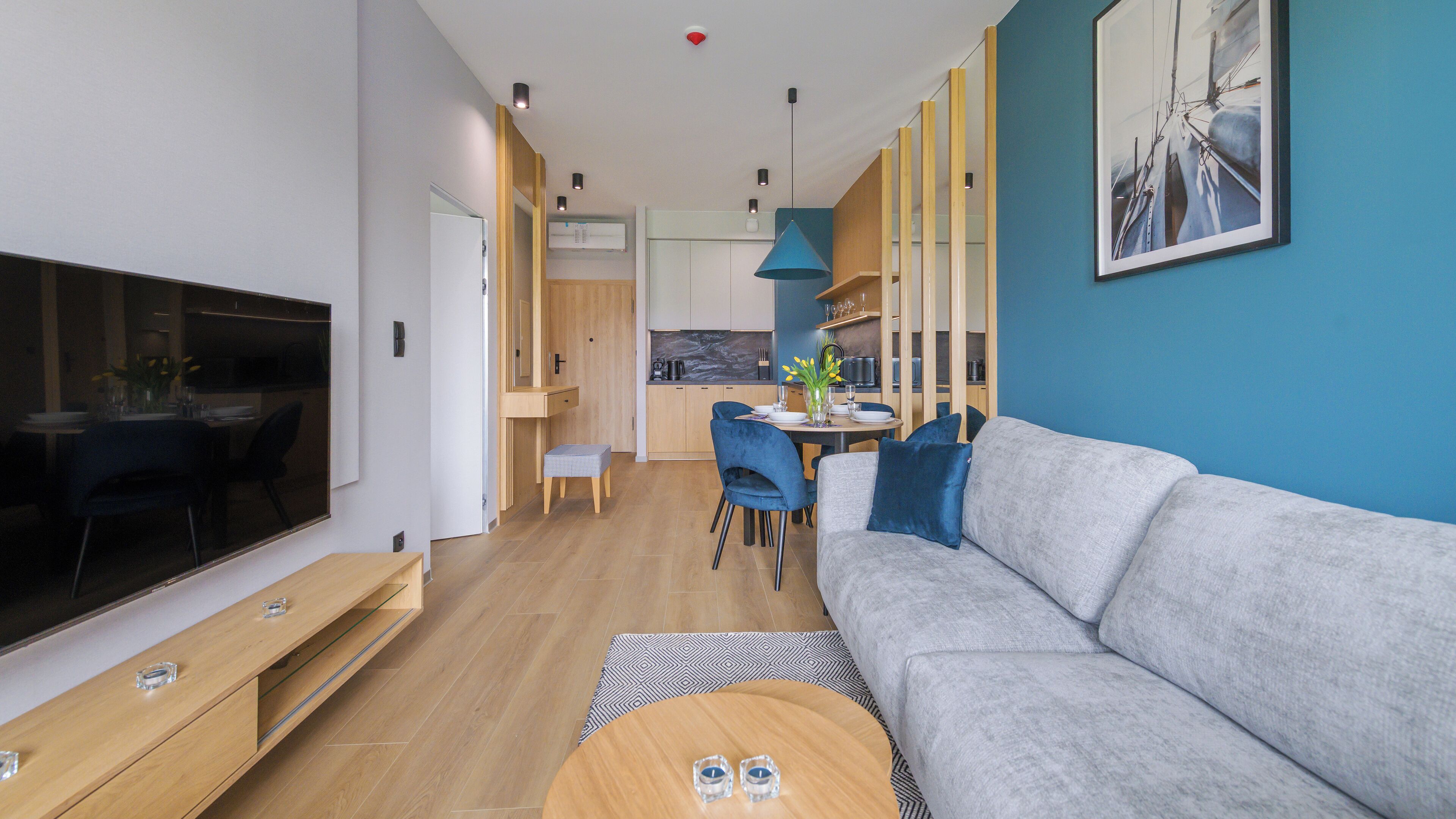 Un salón moderno con un sofá gris, una mesa de centro de madera y una televisión. Hay una zona de comedor con una mesa y sillas. El suelo es de madera y una pared de acento es azul.