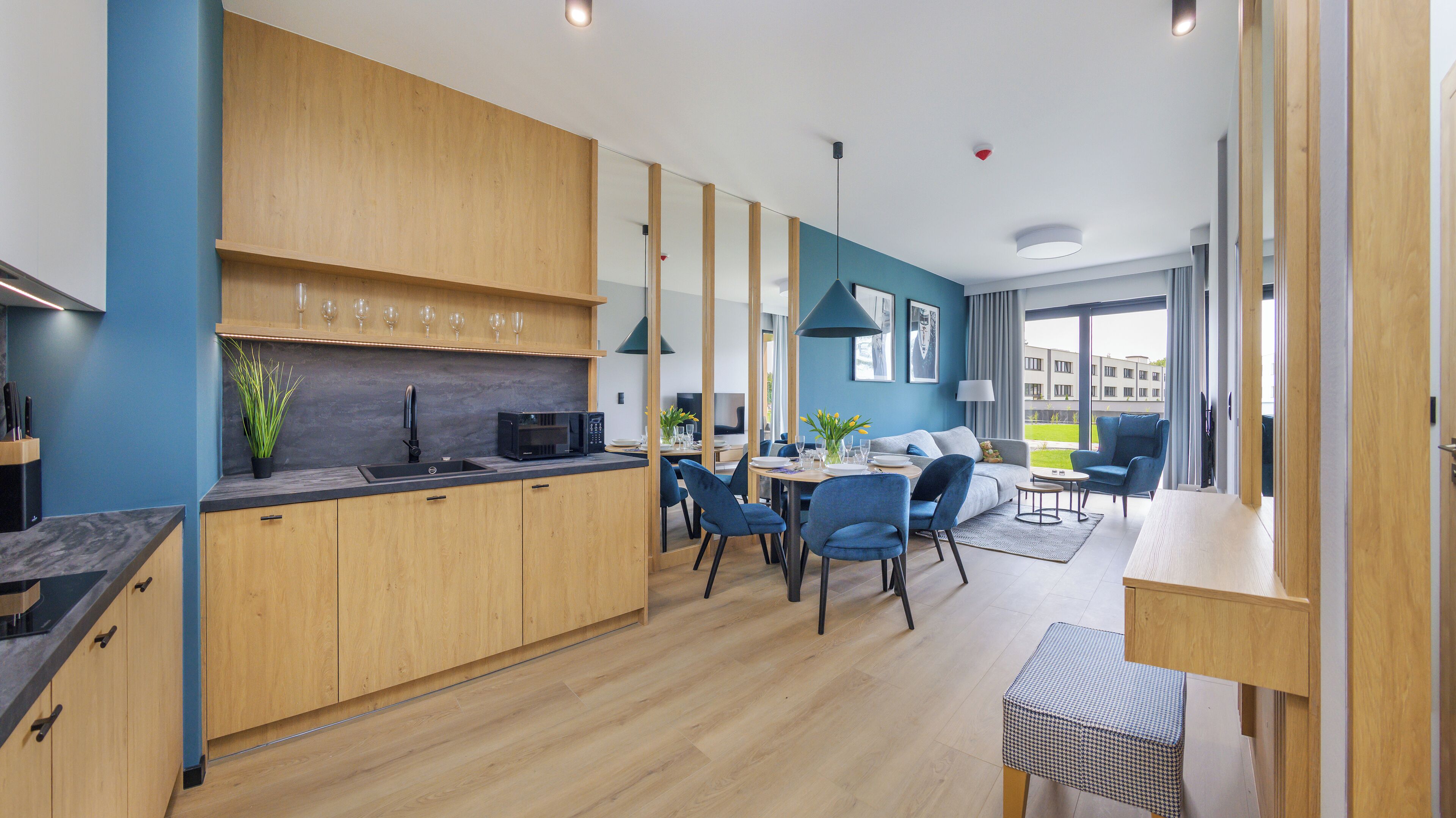 A modern kitchen with wooden cabinets and a built-in microwave. There is a dining area with blue chairs and a small table.