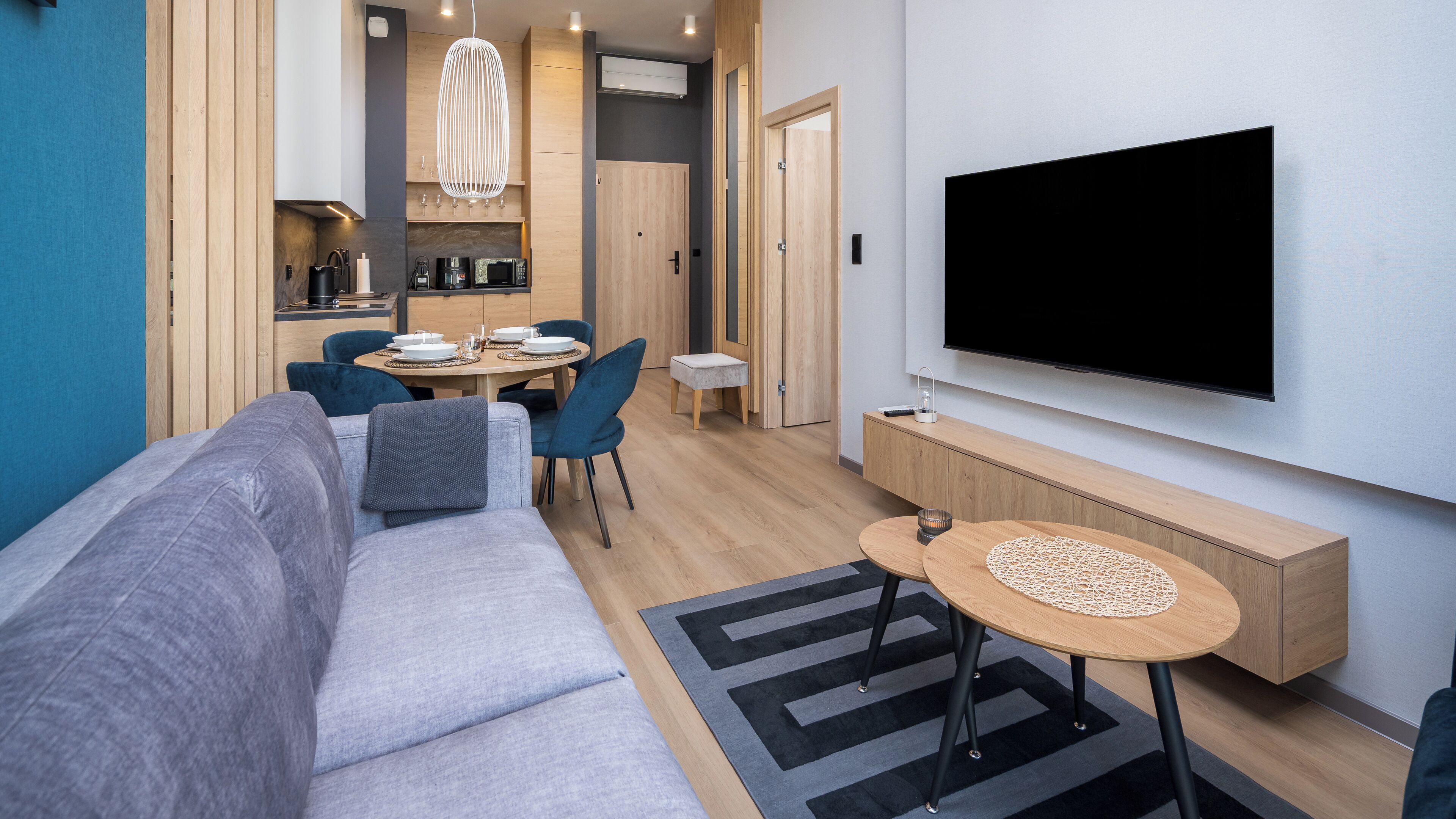 A modern living room with a sofa, dining table, chairs, a TV, and a kitchen area.