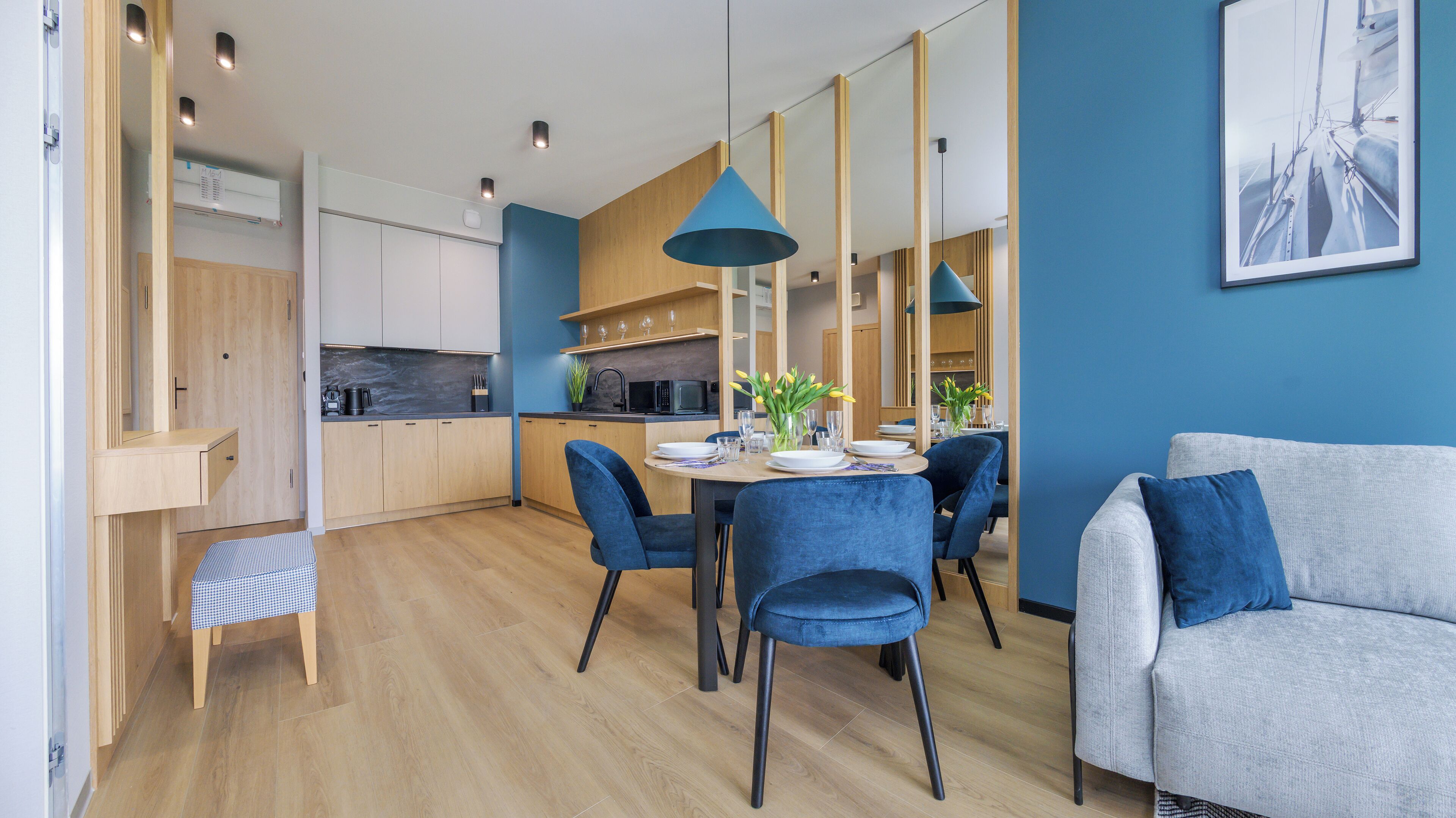 A modern kitchen with wooden cabinets, a dining table with blue chairs, and a grey sofa.