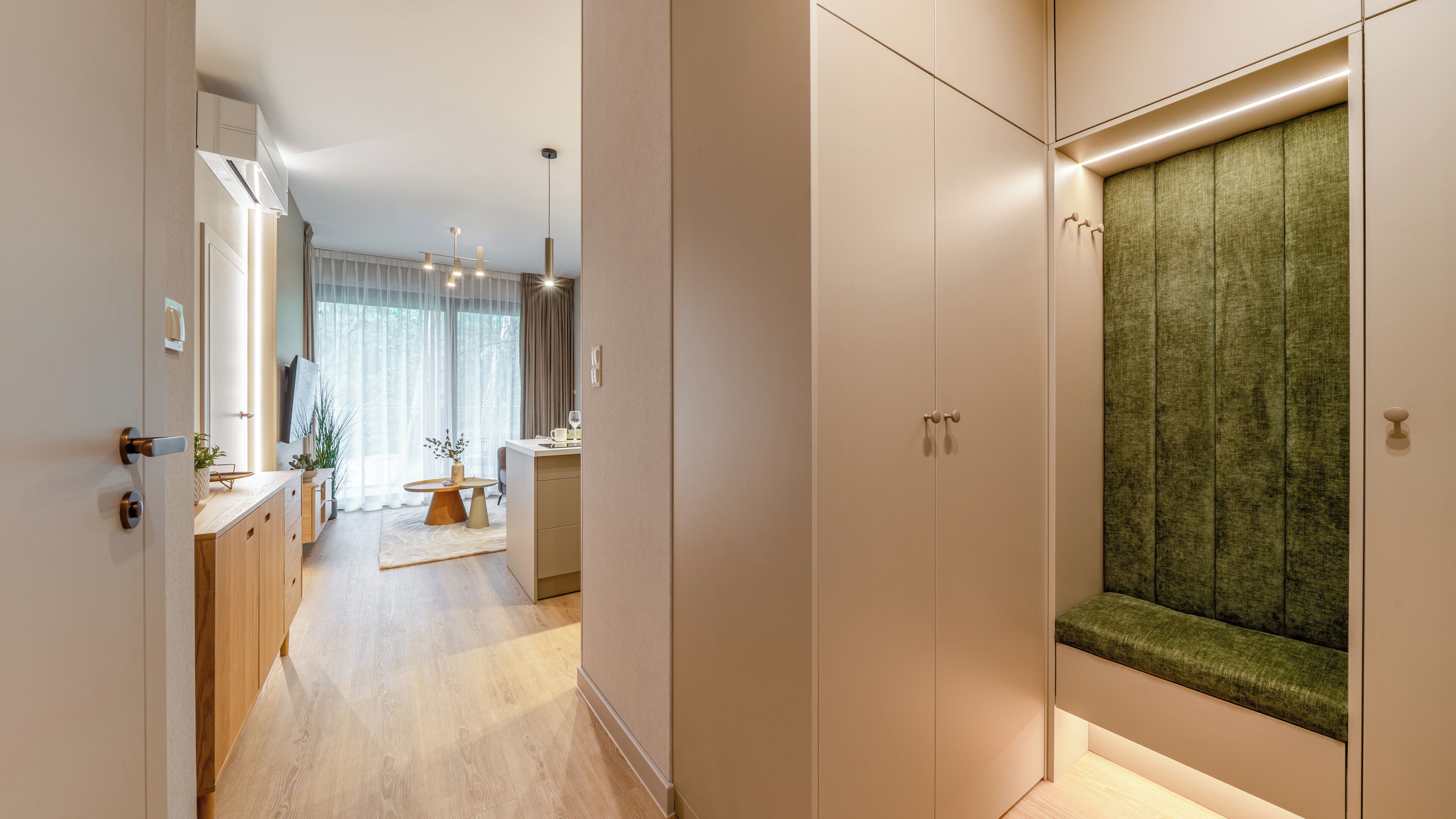 A modern hallway with a wooden floor, a green upholstered bench, and a view into a room with a large window and a dining area.