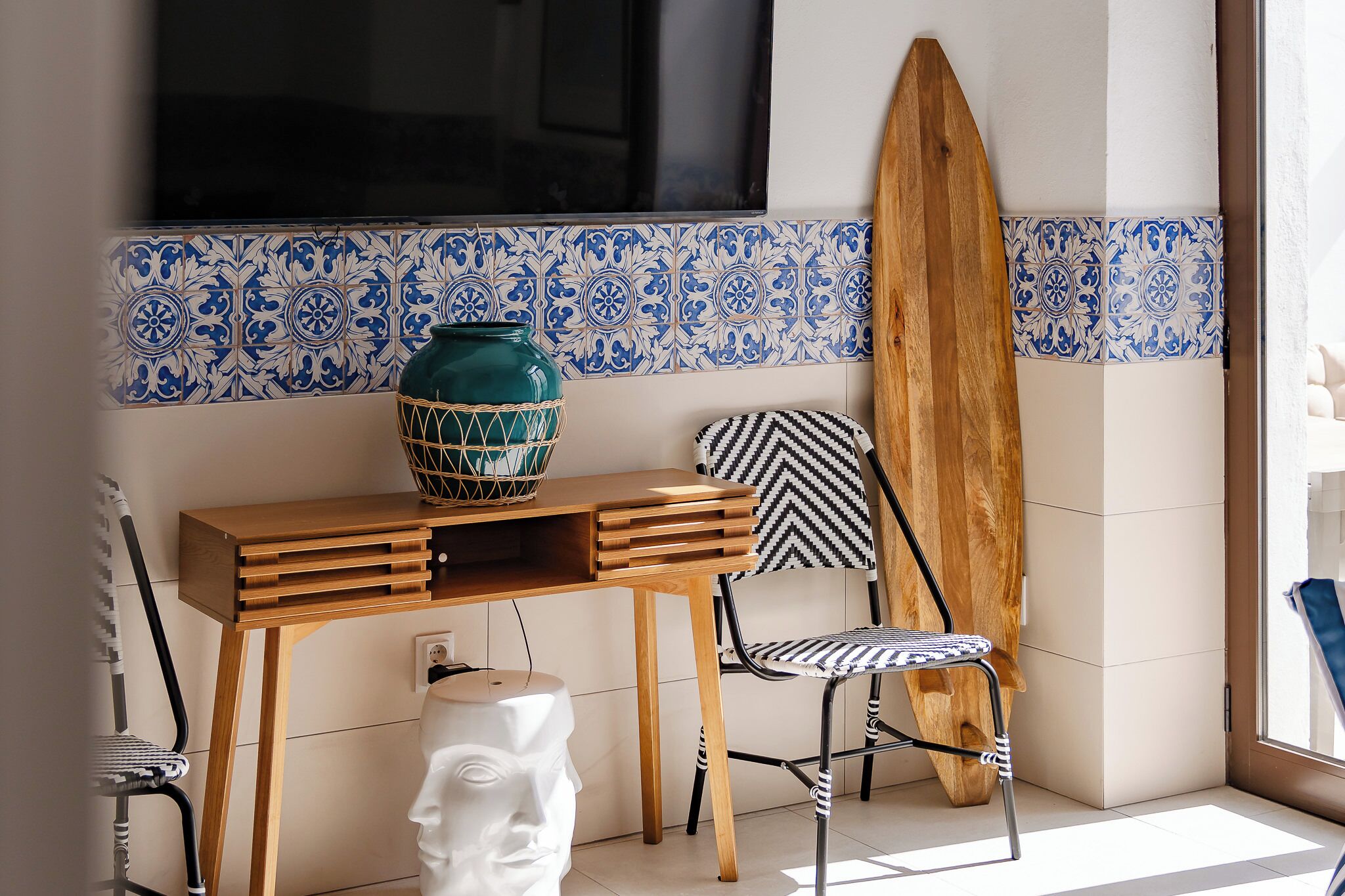 A room with a wooden table, a chair, a surfboard leaning against the wall, and a TV mounted above.