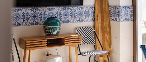 A room with a wooden table, a chair, a surfboard leaning against the wall, and a TV mounted above.