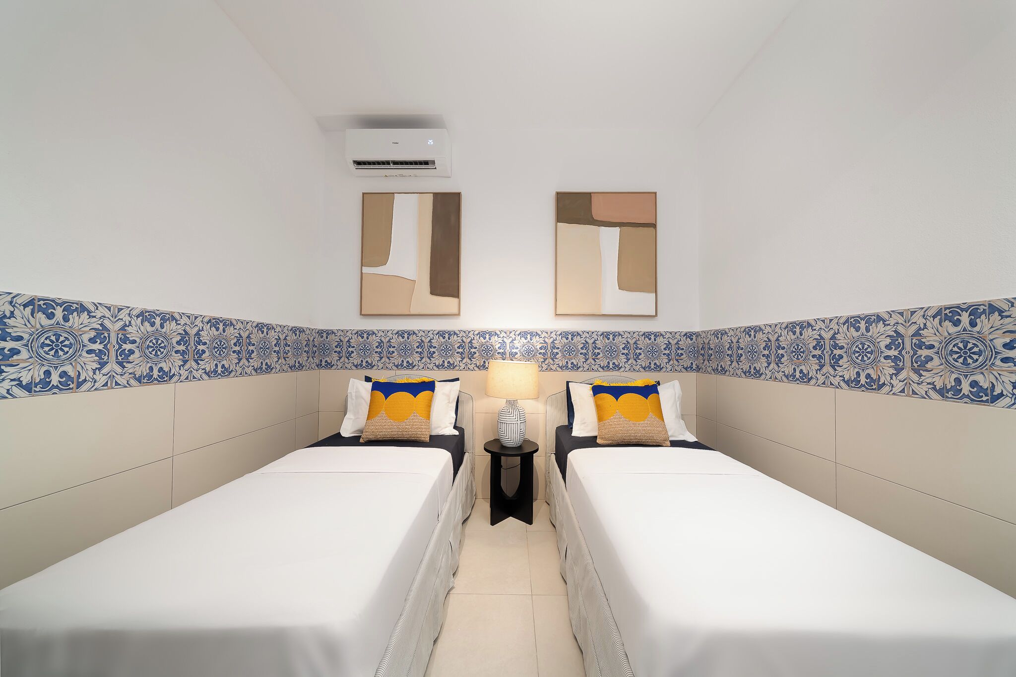 A hotel room with two beds, a nightstand with a lamp, and decorative wall tiles.