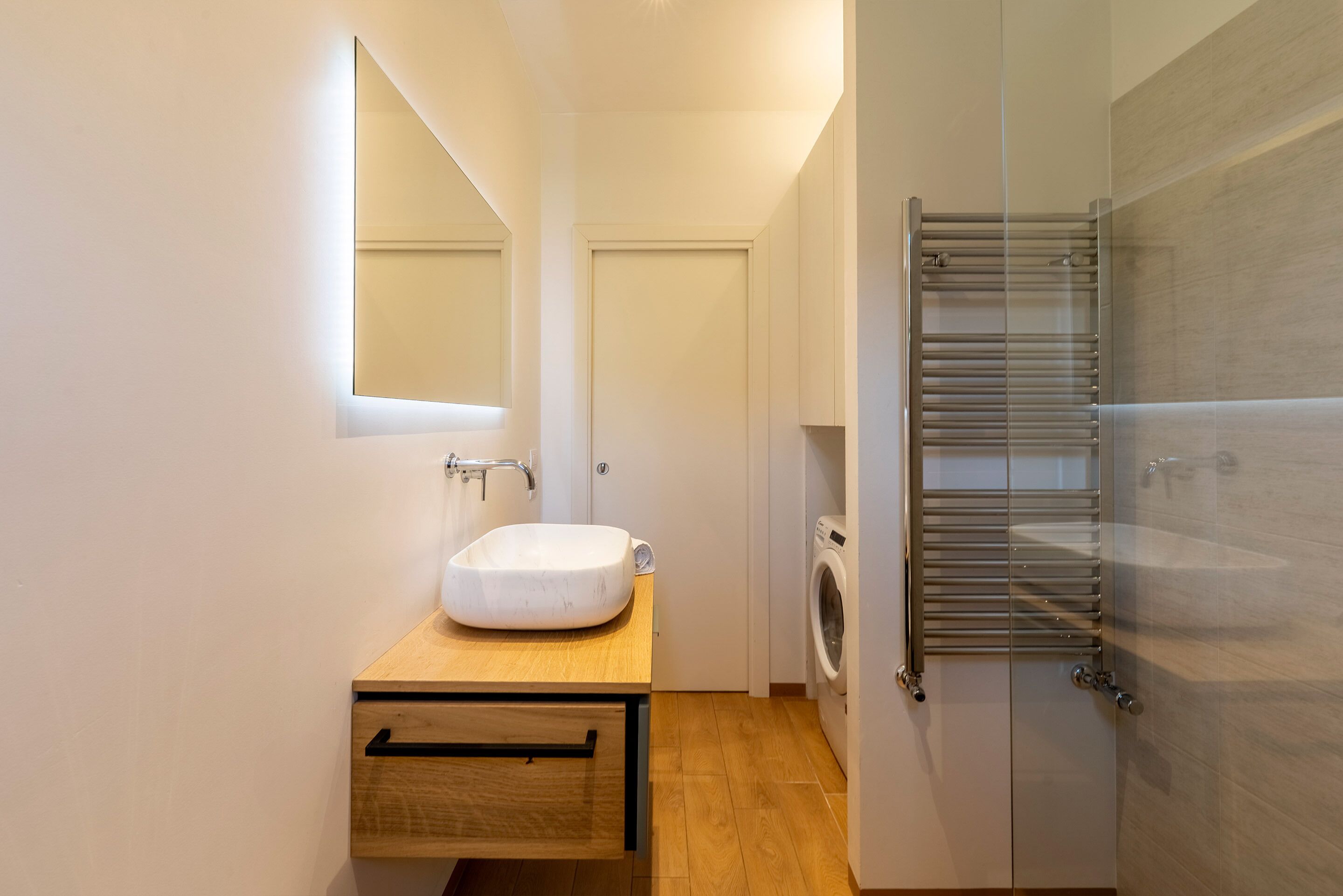 A modern bathroom with a wooden vanity, a white sink, a mirror, a towel rack, and a washing machine.