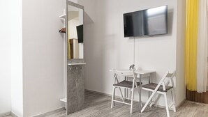 Quarto moderno com televisão de tela plana na parede, uma pequena mesa branca, duas cadeiras brancas e um armário com espelho.