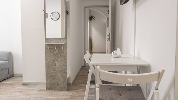 Quarto moderno e minimalista, com uma mesa e cadeiras brancas, um relógio na parede e uma escada que leva a outra área.