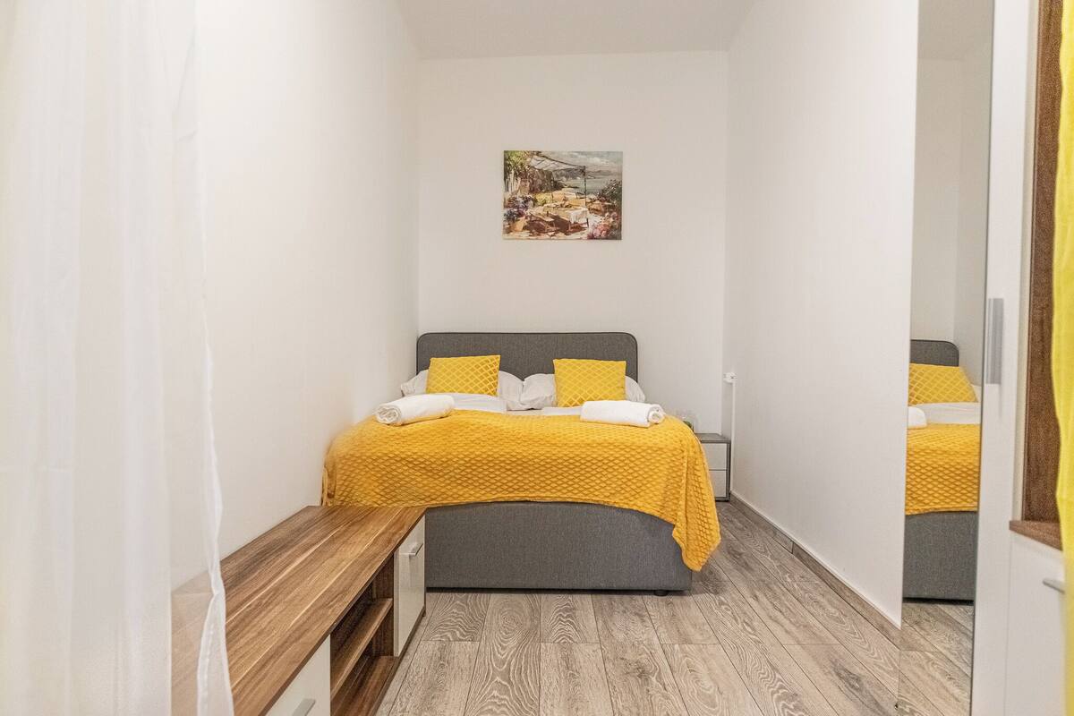 A bedroom with a bed, a bench, a painting on the wall, and a mirror.