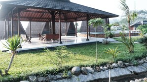 A covered outdoor seating area with a modern design, surrounded by a well-maintained garden with various plants and a stone wall. - Jambu Klutuk Resort (Parakan)