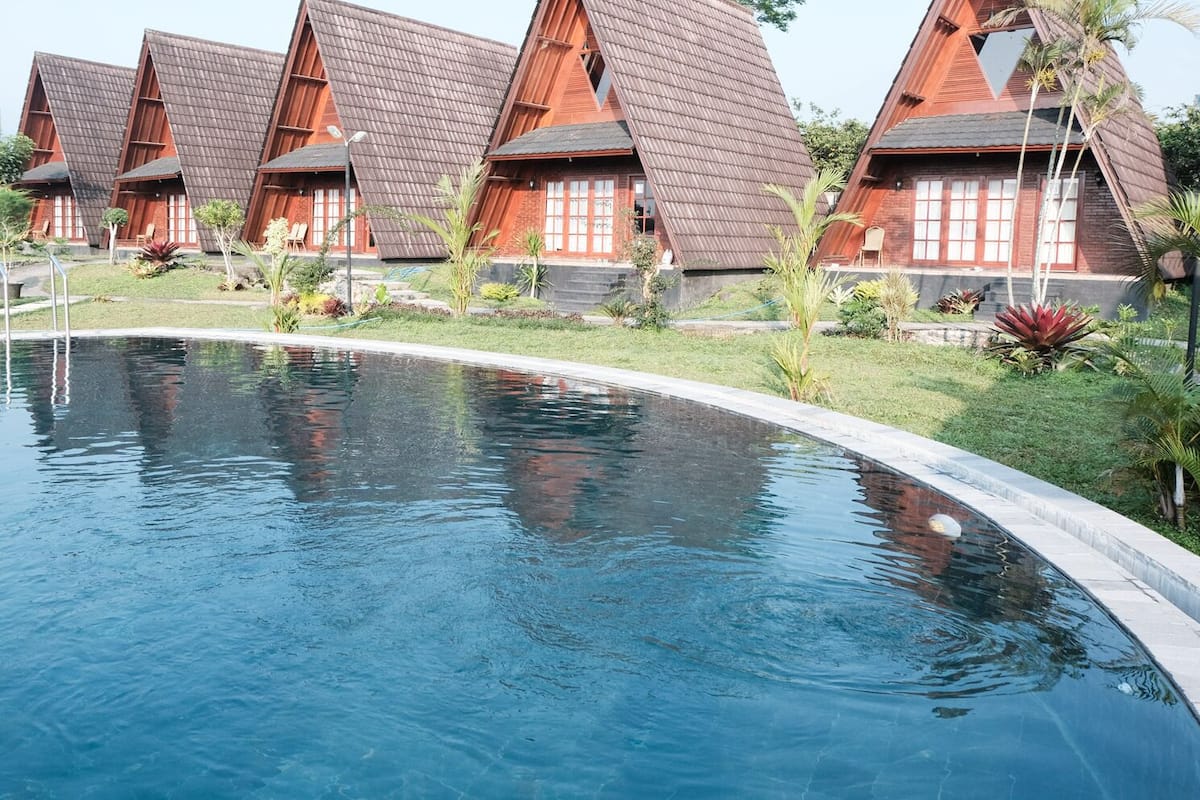 Une piscine à bord incurvé, entourée d’une pelouse bien entretenue et de plantes tropicales. À côté de la piscine se trouvent plusieurs bâtiments en bois dotés de toits inclinés, qui font probablement partie d’un complexe hôtelier ou d’un hôtel.