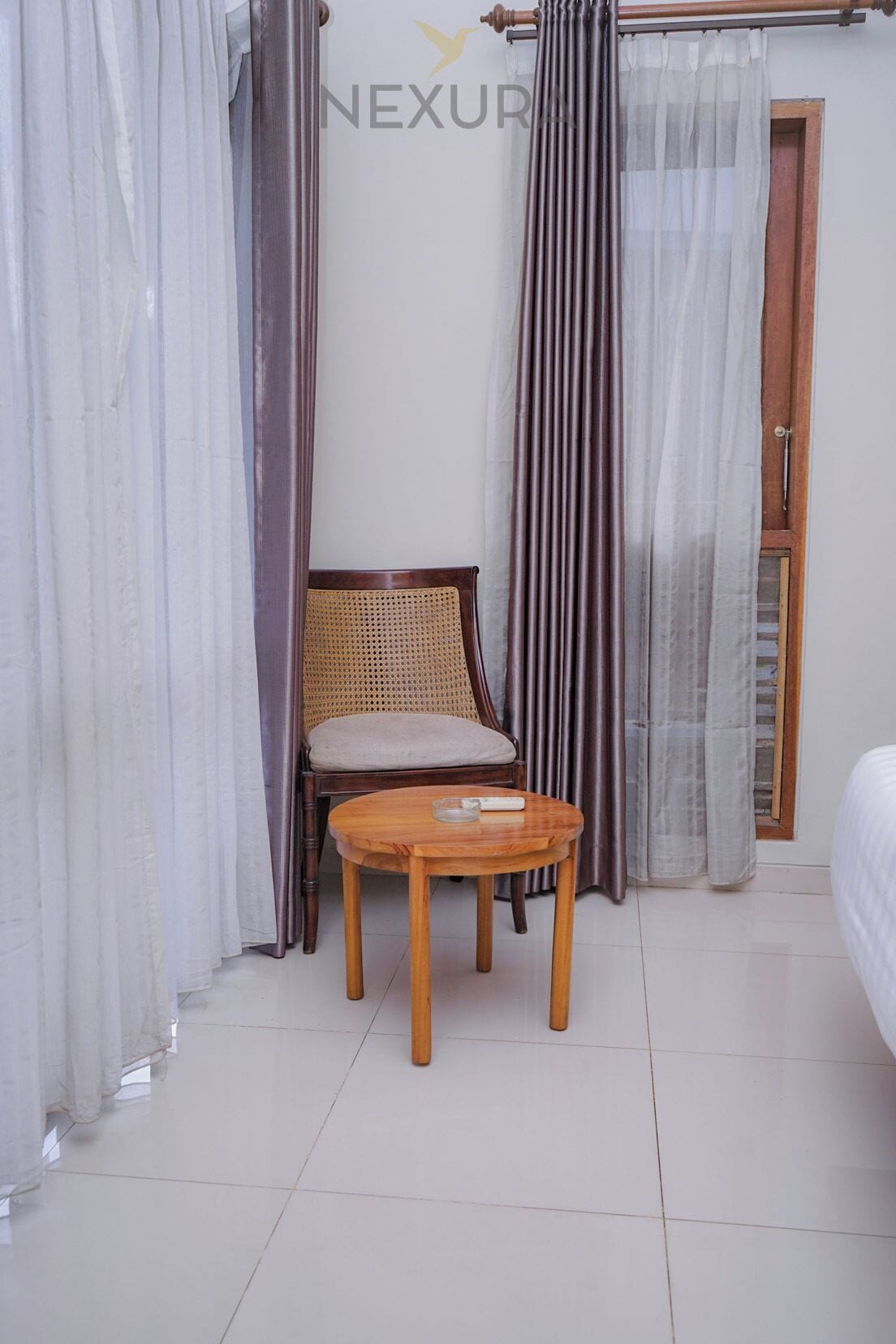 A room with a wooden chair and a small round table, white curtains, and a wooden door.