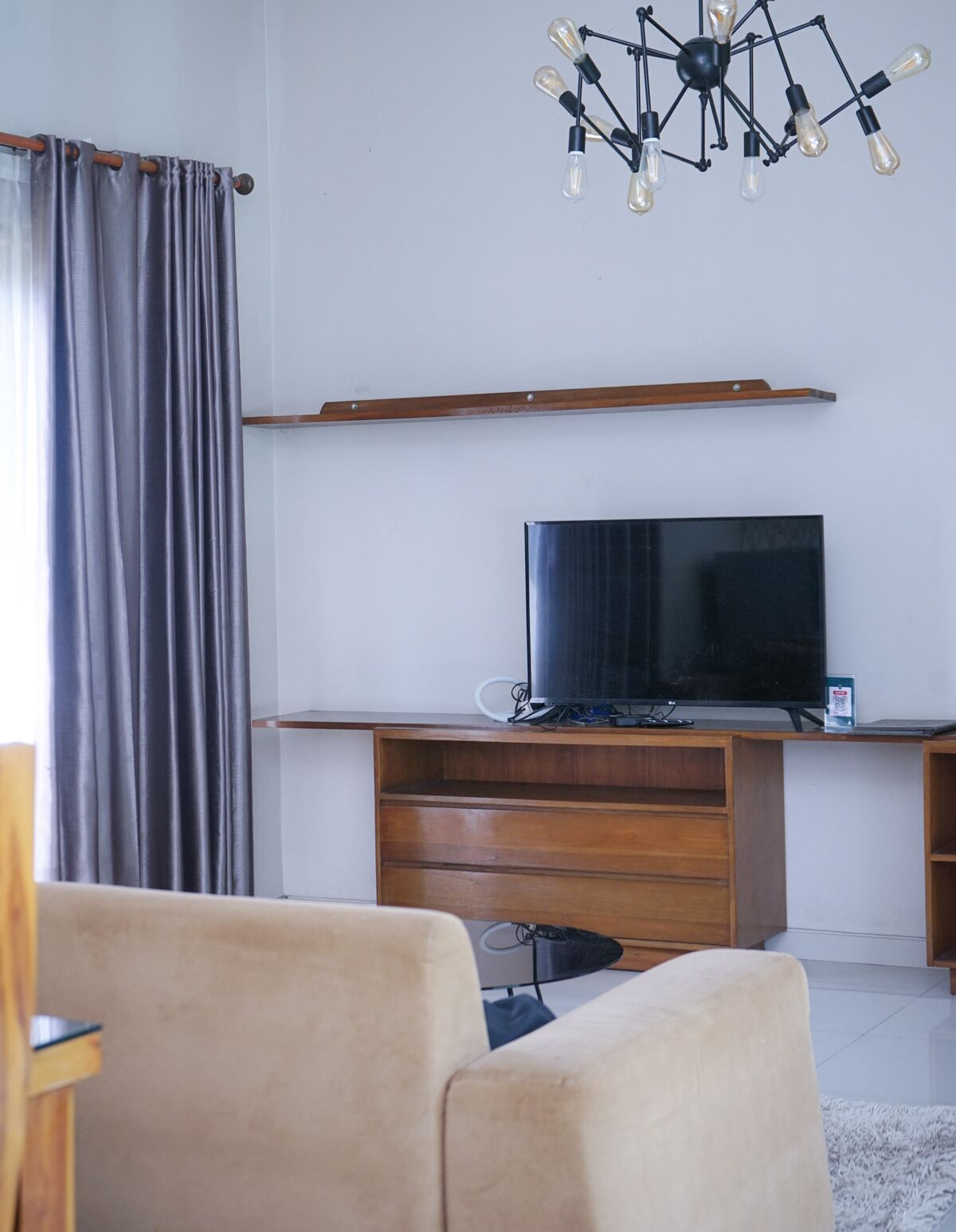 A modern living room with a flat-screen TV on a wooden stand, a chandelier, and a large grey curtain.