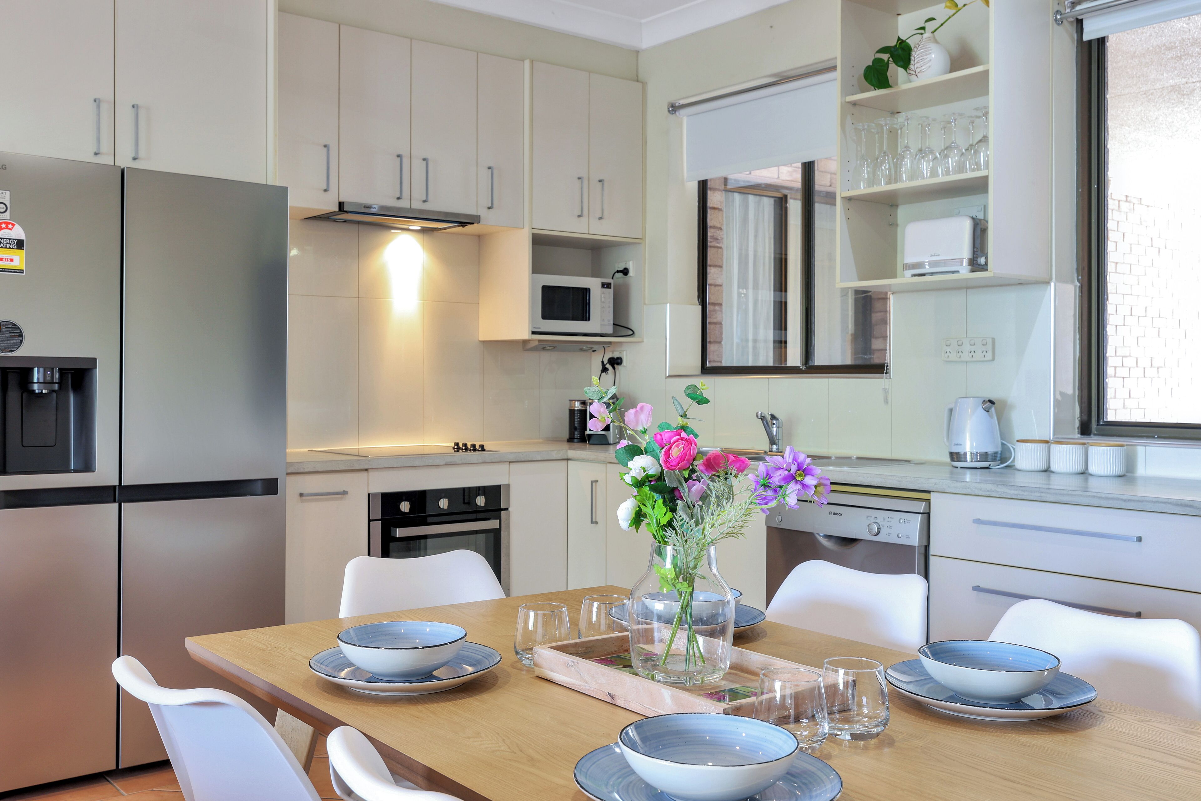 A modern kitchen with white cabinets, a stainless steel refrigerator, a microwave, and a dining table set with bowls and glasses.