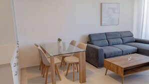 A modern living room with a glass dining table, beige chairs, a grey sofa, and a wooden coffee table. - Phaedrus Living: Seaside Thalassa Cretan D101 (Platanias)
