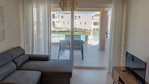 A modern living room with a grey sofa, a TV on a wooden stand, and a view of a pool and apartment buildings through a glass door. - Phaedrus Living: Seaside Thalassa Cretan D101 (Platanias)