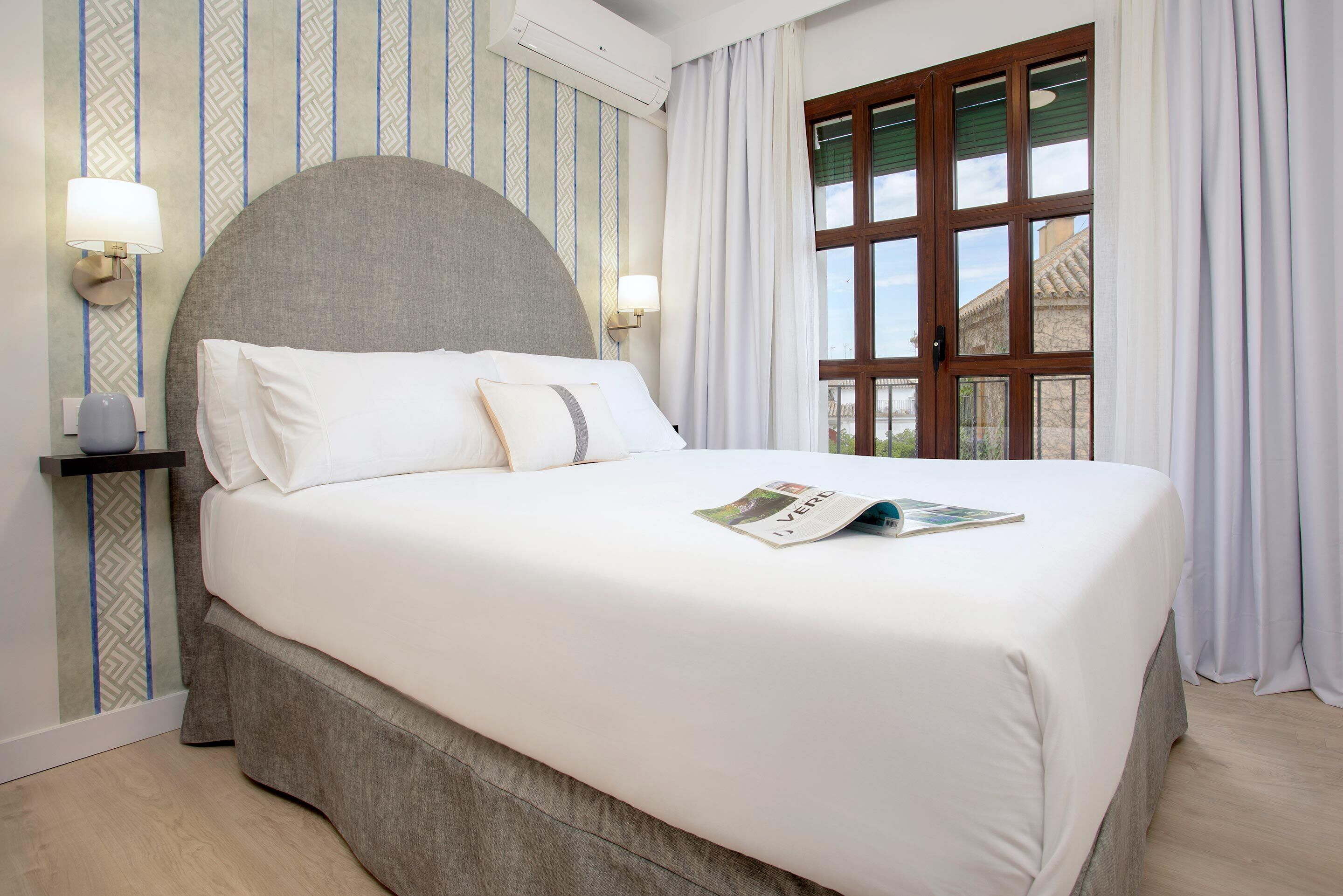 A neatly made bed with white linens, two bedside lamps, and a window with curtains.