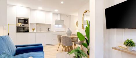 A modern living room with a blue sofa, a dining table with chairs, a kitchen area with white cabinets, and a flat-screen TV mounted on the wall.