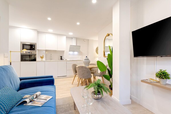 A modern living room with a blue sofa, a dining table with chairs, a kitchen area with white cabinets, and a flat-screen TV mounted on the wall. - Cozy 2bd Apartment in Seville. Conde de Ibarra IV (Seville)
